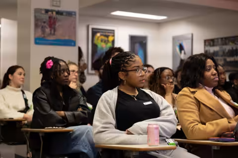 Students are seated at desks in a classroom with posters on the wall and fluorescent lighting overhead. The student in the front center, listening attentively to an instructor out of view, is wearing glasses and a black and gray sweatshirt. 