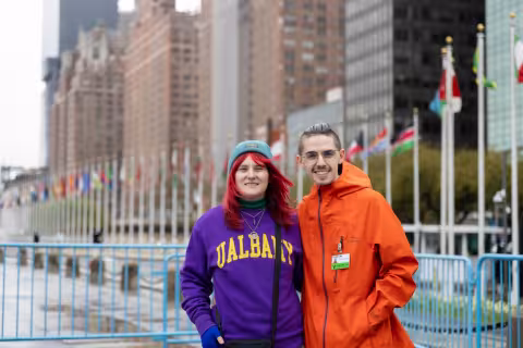 Two people stand in front of a row of flags representing the U.N. member nations. Tall city buildings are in the background, behind the flags. The student on the left has red hair and is wearing a blue hat and purple UAlbany hoodie. The student on the right is wearing a bright orange jacket and glasses. Both are smiling.  