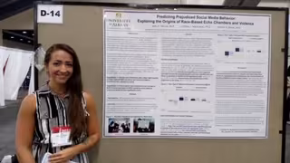 A woman with long brown hair. in a black and white dress wears a name tag and smiles next to a research poster