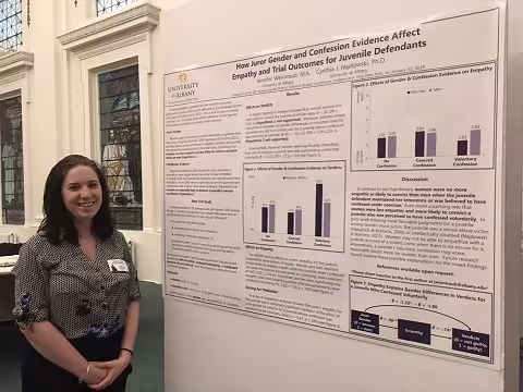 A brunette in a black and white blouse and a name tag smiles and stands next to a research poster mounted on a wall.