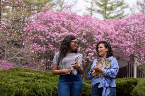 Two women walking outside and talking, with pink spring flowers and greenery behind them.