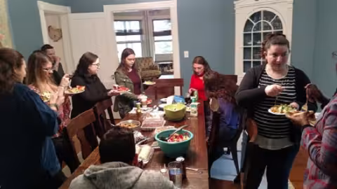 Ten people sit and stand around a dining room table, eating, talking and laughing