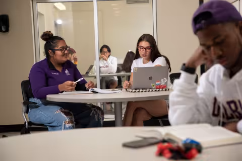Two students sit at a table with a notebook and laptop inside a residence hall study lounge