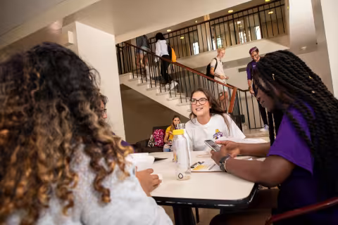 A student smiles as she talks to three other friends seated at a table with her. Other students walk through the residence hall and up its stairs.