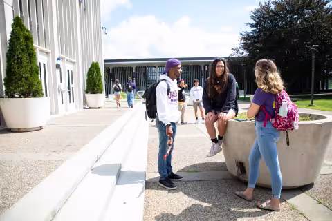 Three students stand outside Eastman Tower talking on a sunny day, while other students walk by.