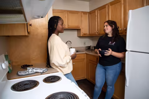 Two students stand in a kitchen holding a mug and bowl, smiling at each other.