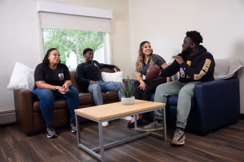 Four students sit on two coaches in a living room with a coffee table and a UAlbany pennant.