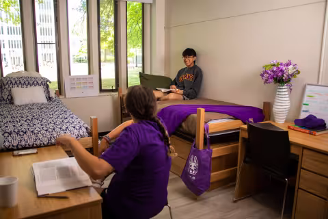 A double bedroom, with two twin beds, two desks and two chairs. One student sits at a desk and another sits on the bed, smiling and laughing together.