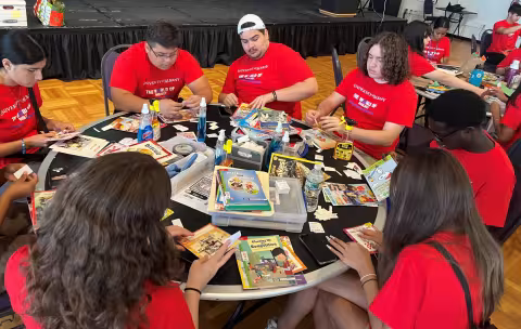 Eight students in matching red T-shirts that say "The World of Biology" are seating around a table and mending used children's books.