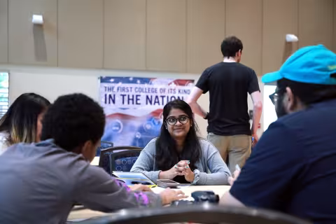 Students participating in a CEHC game design challenge sit facing each other around a circular table with a CEHC banner in the background that reads: "The first of its kind in the nation"