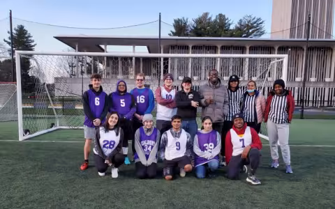 14 members of the UAlbany unified soccer league pose smiling in two rows in front of a soccer goal on the Dutch turf field with Dutch Quad in the background. nights this semester. (