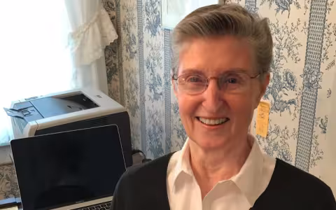 A portrait of Joan Blanchfield at home, smiling and wearing a white collared shirt and dark sweater. 