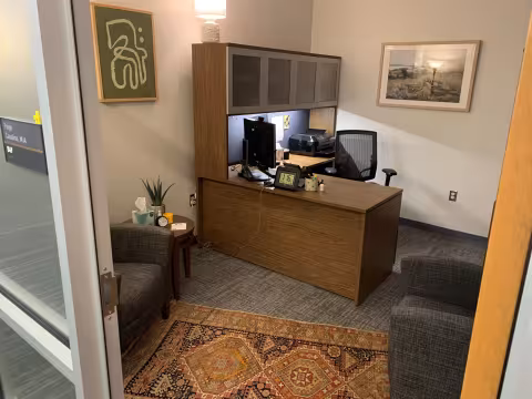 The view into an office with an L-shaped desk and two arm chairs facing each other. There is art on the walls and a patterned carpet on the ground.