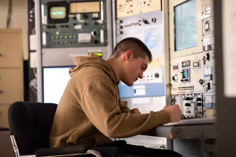 A student sits at a desk in front of an instrument panel and writes in a notebook.