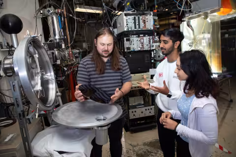 An instructor holds a circular mirror in his hands and stands over an open piece of equipment, as two students look on.