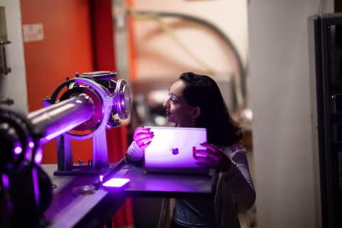 A student stands in front of a piece of equipment, looking in a small window while holding an iPad in front of herself.