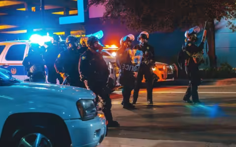 Police in riot gear stand amid cars on a street lit by headlights