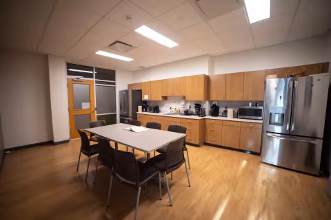 A kitchen with two large silver fridges, a microwave, several coffee makers, a sink, light brown cabinets and an eight-seater table.