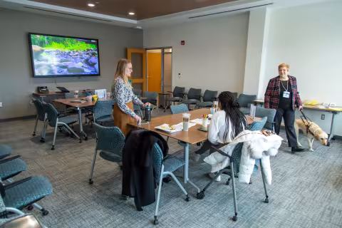 Three women, one of whom has a service dog, speak inside a room with two large tables and many chairs.