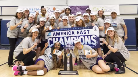 The Great Danes women's basketball team poses on the court next to the America East championship trophy holding a sign declaring them the 2022 America East champions.