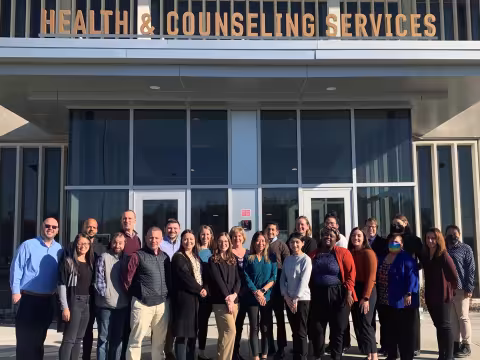 23 CAPS staff members pose in two lines in front of the Health & Counseling Services building.