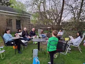 Seven people sit in chairs around a fire pit in a backyard, surrounded by trees and a house, with a child standing with his back facing the camera.