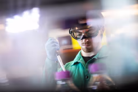 A student with safety googles, a green lab coat and blue gloves uses an instrument to measure a sample in a biology lab. He is seen through other instrumentation and equipment.