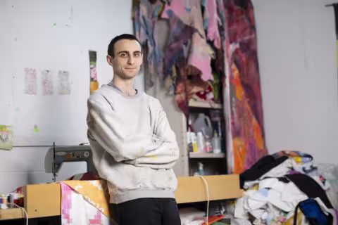 A man with dark hair wears a sweater inside out and leans against a desk in his studio, arms folded. A tall shelf in the background is covered in towels and fabric. A sewing machine sits on the desk behind him. 