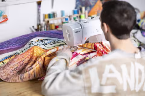 A man with dark hair uses a sewing machine on a colorful towel. Spools of thread are pictured in the background.