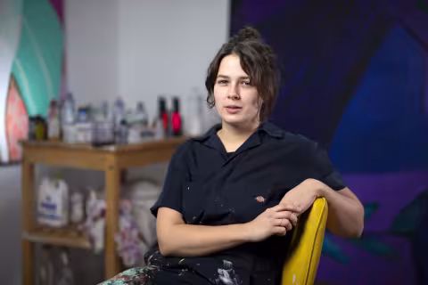 A woman with dark brown hair pulled into a claw clip wears a navy, paint-splattered jumpsuit and poses for a portrait in a yellow chair. In the background are two large paintings and a wooden cart holding paint supplies.