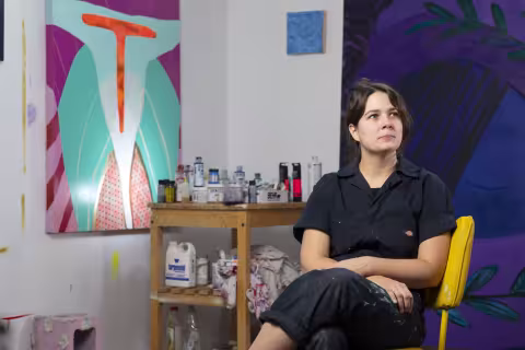 A woman with dark brown hair pulled into a claw clip wears a navy, paint-splattered jumpsuit and looks into the distance as she poses for a portrait in a yellow chair. In the background are two large colorful paintings and a small blue painting, a wooden cart holding paint supplies, and a pink cinder block.