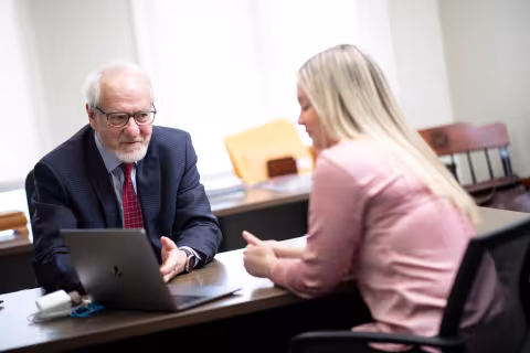 A professor and a student sit across from each other looking over a laptop in an office with a bookshelf in the background.