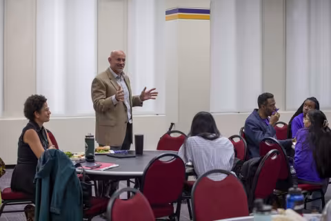 Paul Morgan, standing at a table in a room in the campus center, welcomes the SPRINTER students. 