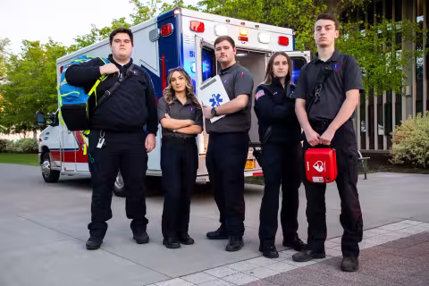 Five ambulance crew members in Five Quad uniforms and holding gear pose for a photo in front of an ambulance. 