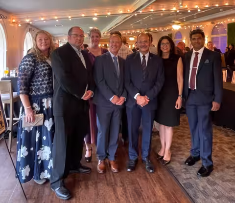 Seven people representing University at Albany leadership and the RNA Institute stand in a row, smiling for a photo at the Muscular Dystrophy Association event. Festive lighting borders the image. Left to right: Tammy Reid, John Cleary, Sheila Seery, Andy Berglund, President Havidán Rodríguez, Rosy Rodríguez, Jibin Abraham Punnoose. 