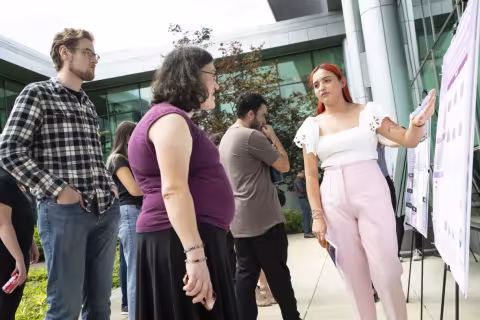 A young woman wearing a white short-sleeved blouse and pink pants stands, smiling, next to her research poster. The photo was taken outdoors in the life sciences building courtyard on a sunny day. The student is presenting her work to interested onlookers.  