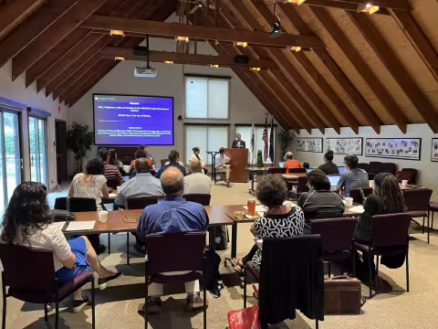  Meeting attendees face the front of the room as a PowerPoint presentation is delivered by a speaker at a podium.