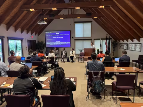 Meeting attendees face the front of the room as a PowerPoint presentation is delivered. Three panelists are seated at the front of the room, with one speaker standing at the podium. 