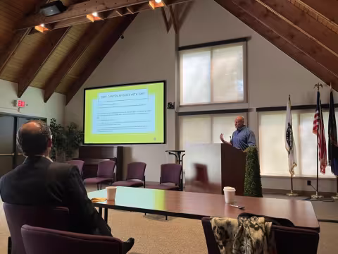  Paul Morgan at a podium delivering a presentation. A lime green PowerPoint side to his left reads: “How can you affiliate with ISHE?”. The acronym “ISHE” stands for Institute of Social and Health Equity. 
