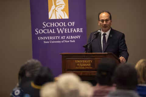 President Havidán Rodríguez addresses the audience, standing in front of a purple banner which says "School of Social Welfare". He is wearing glasses, a dark suit jacket and purple tie.