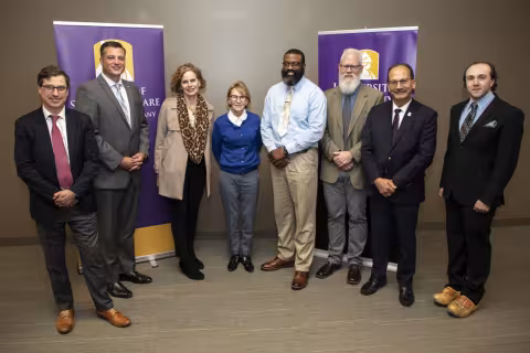 The event's eight speakers stand in front of two purple banners, one which says "School of Social Welfare," the other says "University at Albany". The speakers are standing against a tan wall with tan carpeting on the floor.