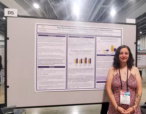 A woman in a red and white floral dress, wearing a lanyard with shoulder-length brown hair stands in front of a research poster titled, 'Raced-Based Difference in Perceptions of Freedom in Police Encounters'