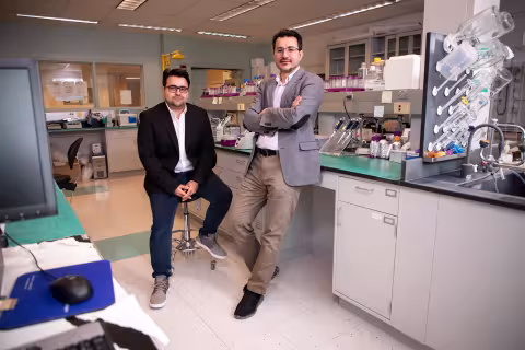 Two men pose for a portrait in a chemistry lab. One is seated, wearing a black blazer and jeans; the other is leaning against a lab bench wearing a gray blazer and khaki pants. Various glass and plastic containers are on the bench and shelf behind them.