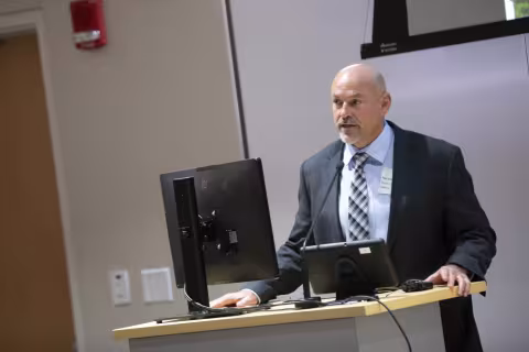 A man wearing a dark gray suit and plaid tie stands at a podium in UAlbany's ETEC building to address an audience. 
