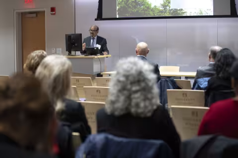 A man wearing dark rimmed glasses, striped tie and a dark gray suit stands at a podium in UAlbany's ETEC building to address an audience of seated listeners. 