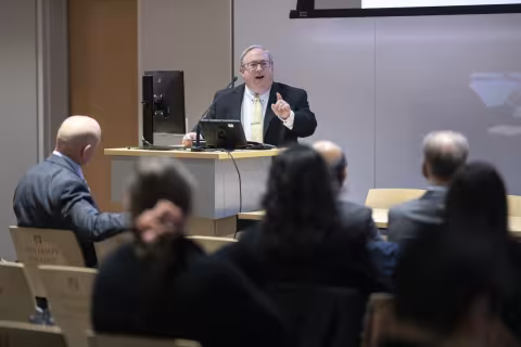 A man wearing square framed glasses, a dark gray suit and pale yellow tie stands at a podium in UAlbany's ETEC building to address an audience of seated listeners. 
