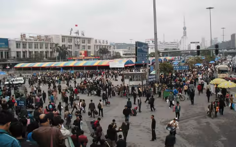 A crowd of people walk in front of Guangzhou Station in China. The station has red, yellow and teal striped awnings out front, a clock on its facade and three signs in Chinese that translate to:  "Unify the motherland," "Guangzhou Station" and "Revitalize China."