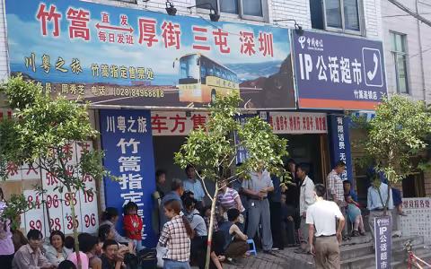 People stand on a street inside outside a storefront adorned with Chinese advertisements for a bus and transportation service.