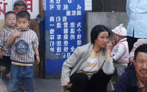 A woman crouches on a stoop in front of a blue and white sign. Children are standing beside her as she sits with her cheek resting on her hand.