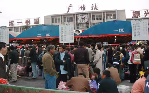 A crowd of people walk in front of Guangzhou Station in China. The station has three red and teal awnings out front, a clock on its facade and signs in Chinese that translate to, "Unify the motherland," "Guangzhou Station" and "Revitalize...," with the remaining text out of frame.